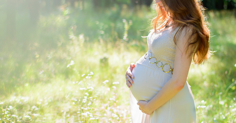 Schwangere Frau im Freien, die einen fließenden, pastellfarbenen Kleid trägt und sanft ihren Babybauch hält.