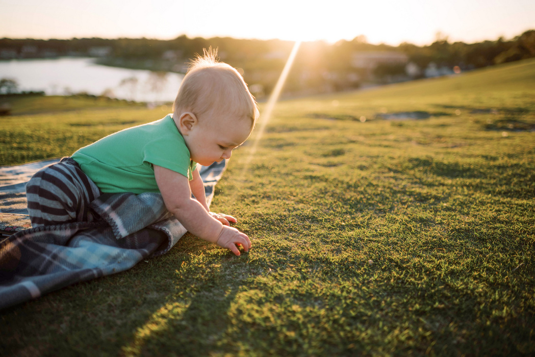 Baby in grünem Shirt liegt auf einer Decke im Gras, bei Sonnenuntergang im Freien.