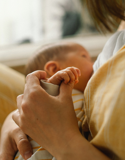Mutter hält sanft die Hand eines Neugeborenen beim Stillen in gelbem Outfit.