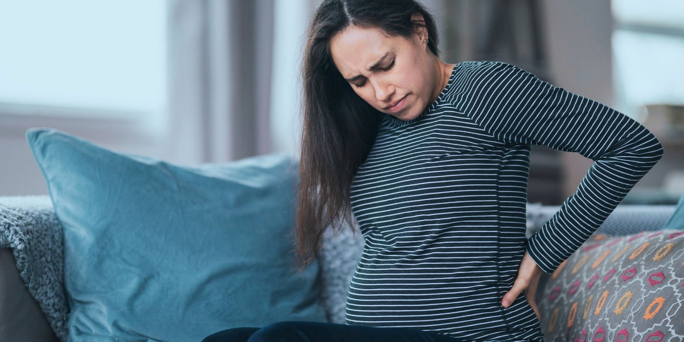 Schwangere Frau sitzt auf einem Sofa und hält ihren schmerzenden Rücken.