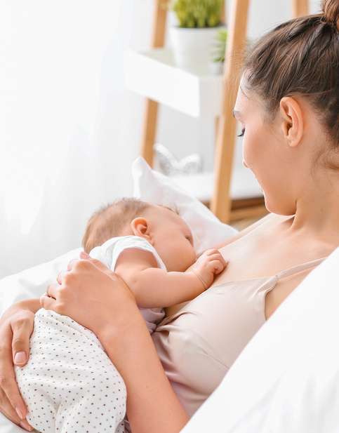 Frau stillt Baby in einem gemütlichen Schlafzimmer, umgeben von weißen Kissen.