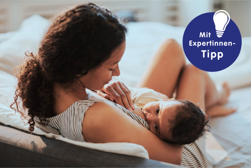 Stillende Mutter hält ihr Baby auf dem Schoß, beide entspannen sich in einem hellen Raum. Experten-Tipp im Bild.