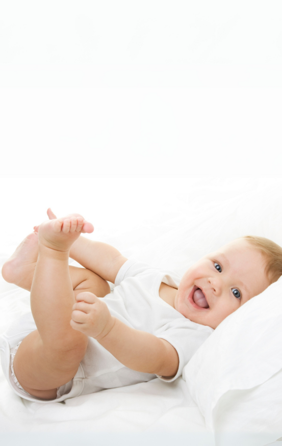 Lächelndes Baby in weißem Strampler liegt auf einem Bett und spielt mit den Füßen.