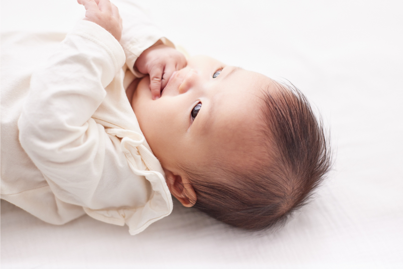 Lächelndes Baby mit dunklen Haaren, das auf einem weißen Untergrund liegt und den Daumen im Mund hat.