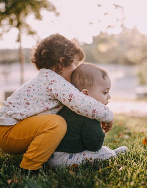 Kleinkind umarmt sitzendes Baby auf einer Wiese im Park bei Sonnenuntergang.
