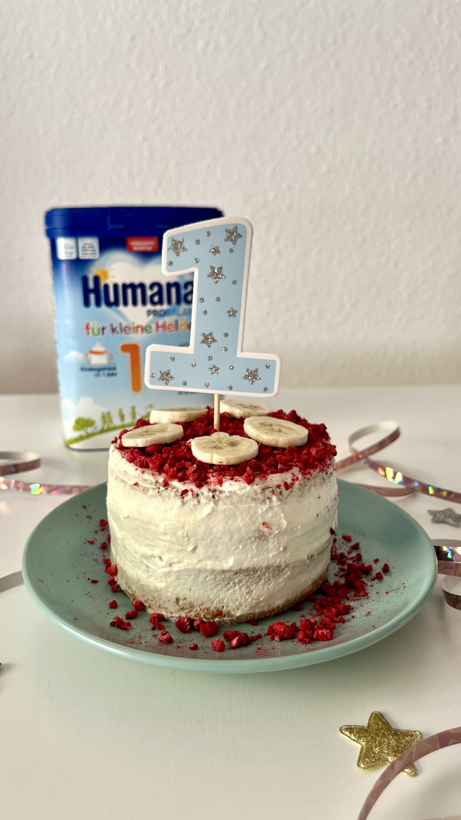 Kleine Torte mit einer blauen „1“-Kerze, dekoriert mit Bananenscheiben und roten Streuseln, auf einem grünen Teller. Im Hintergrund eine Verpackung von Humana für kleine Helden.