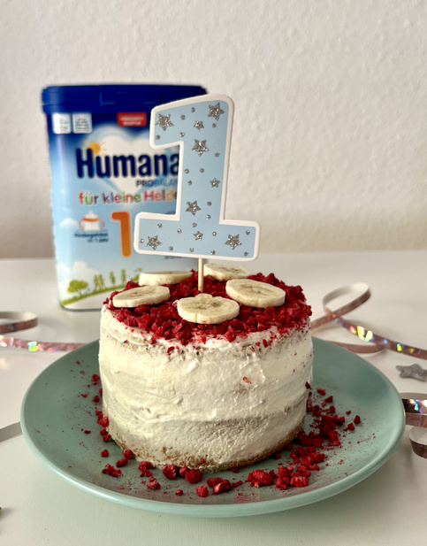 Kleine Torte mit einer blauen „1“-Kerze, dekoriert mit Bananenscheiben und roten Streuseln, auf einem grünen Teller. Im Hintergrund eine Verpackung von Humana für kleine Helden.