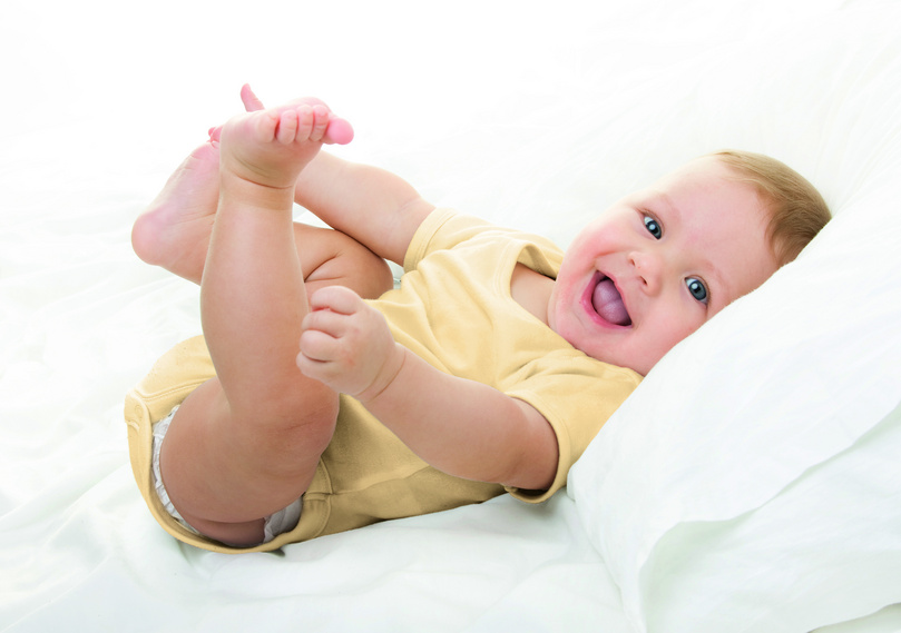 Lächelndes Baby in gelbem Strampler liegt fröhlich auf einem weißen Bett.