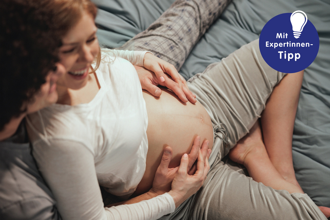 Händchen haltendes Paar entspannt auf einem Bett, die Schwangere umfasst sanft ihren Bauch, lächelnd und glücklich.