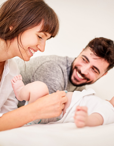 Lächelnde Eltern spielen mit Baby auf Bett. Baby liegt auf dem Rücken und blickt nach oben.