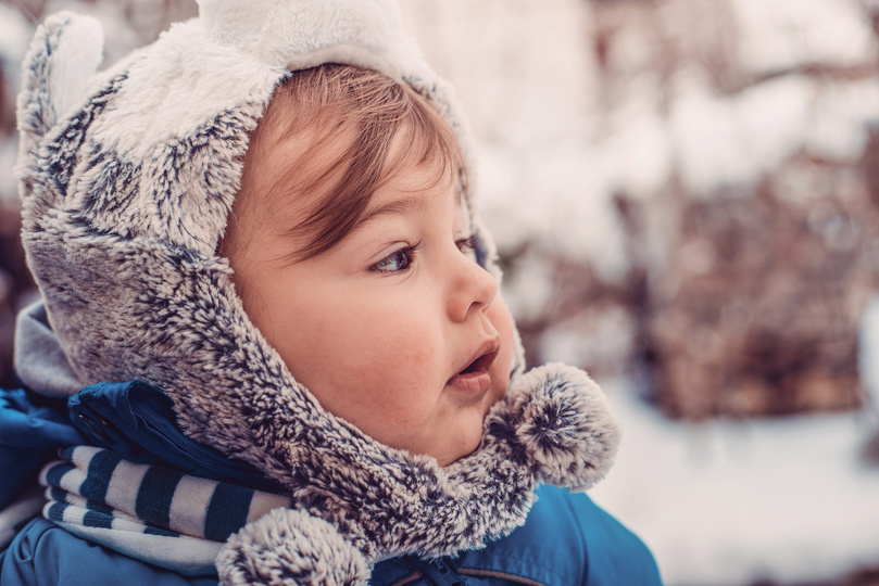 Kleinkind in blauer Winterjacke mit flauschiger Mütze schaut seitlich in eine schneebedeckte Landschaft.