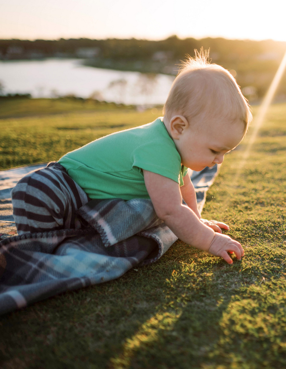 Ein Baby in einem grünen Shirt krabbelt auf einer Decke im Gras bei Sonnenuntergang, nahe einem See.
