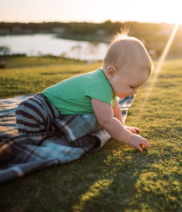 Ein Baby in einem grünen Shirt krabbelt auf einer Decke im Gras bei Sonnenuntergang, nahe einem See.