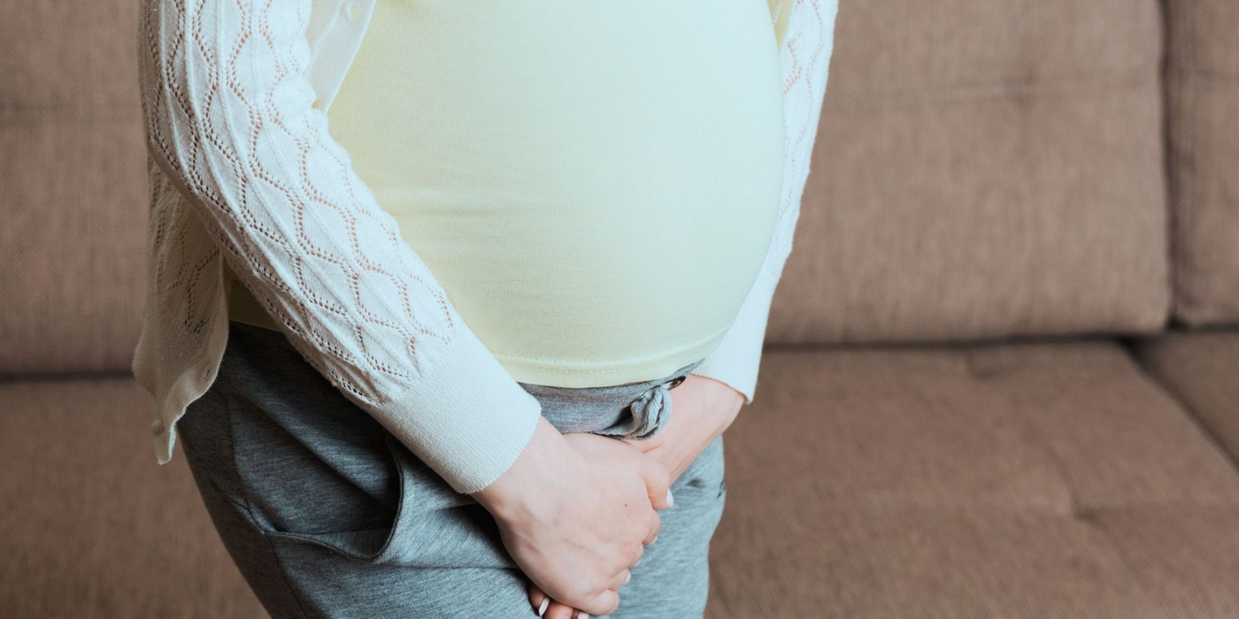 Schwangere Frau hält ihren Bauch, im Hintergrund ein Sofa.