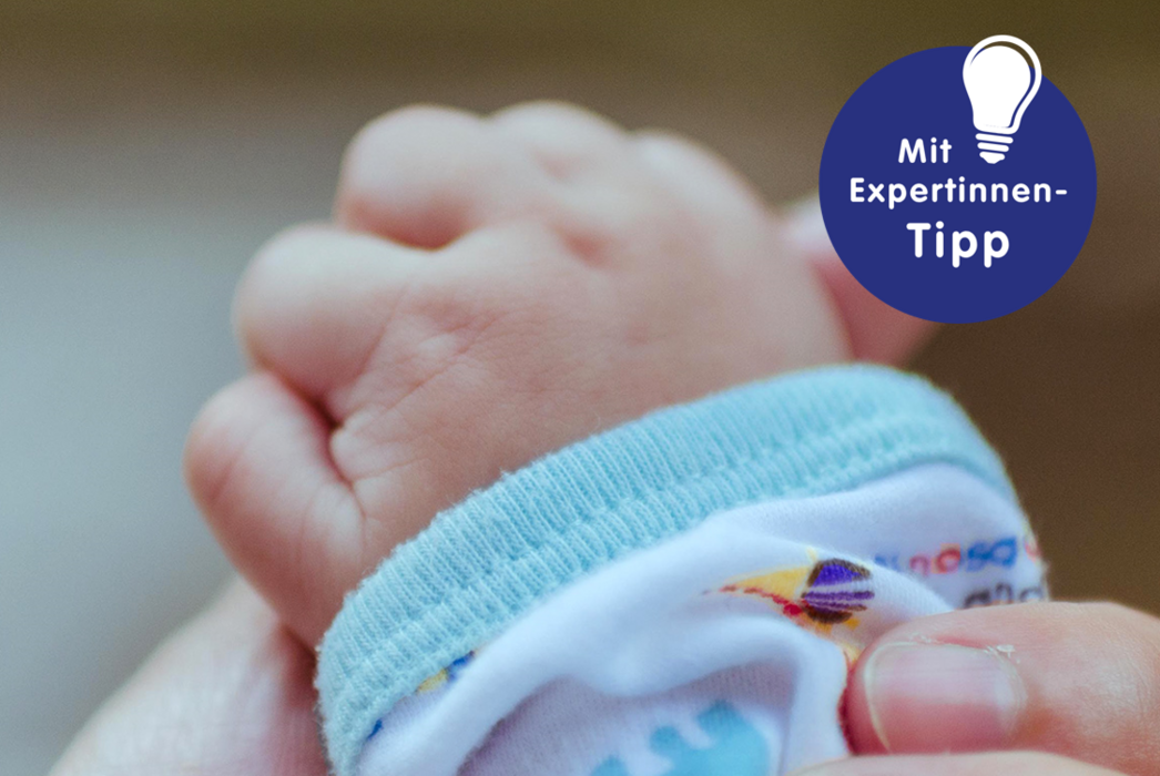 Nahaufnahme einer kleinen Babyhand, die leicht geballt ist und einen farbenfrohen, gemusterten Ärmel trägt, mit einem Experten-Tipp im Eck des Bildes.