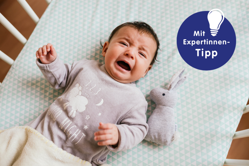 Weinendes Baby in einem Kinderbett, trägt graues Schlafanzug mit Schaf- und Sternenmuster. Neben ihm ein gestreifter Plüschhase.