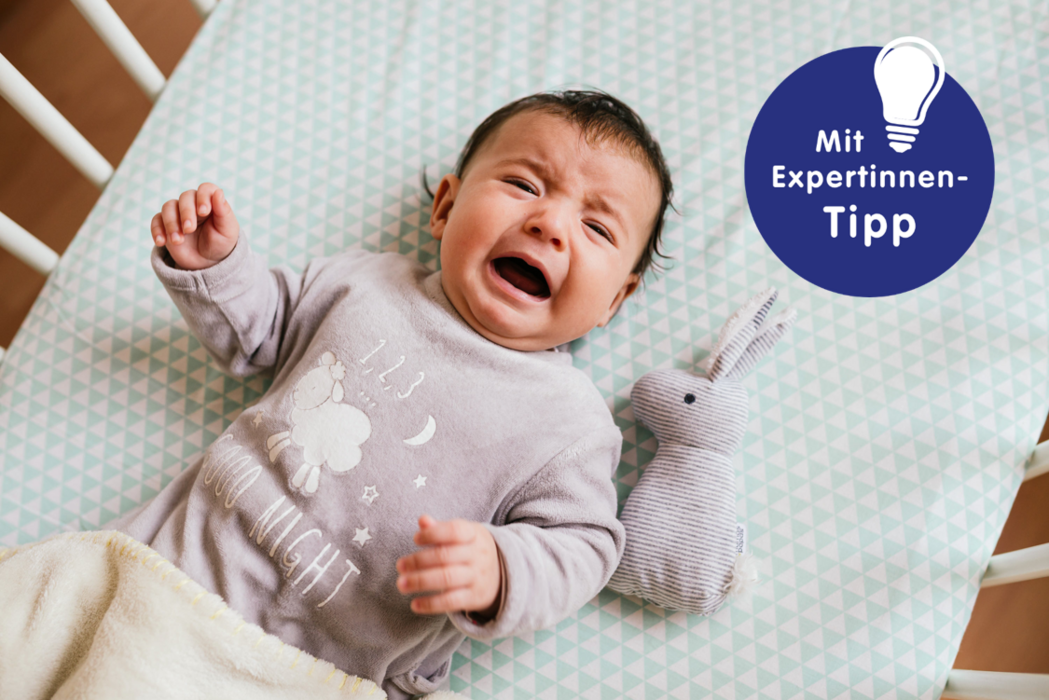 Weinendes Baby in einem Kinderbett, trägt graues Schlafanzug mit Schaf- und Sternenmuster. Neben ihm ein gestreifter Plüschhase.