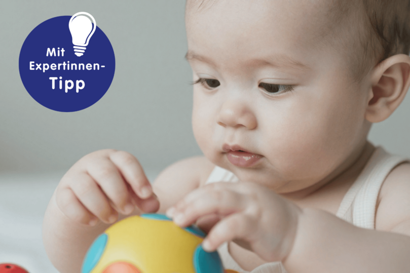 Ein Baby spielt aufmerksam mit einem bunten Ballspielzeug, während im Hintergrund ein Experten-Tipp eingeblendet ist.