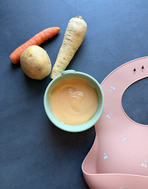 Gemüsepüree in einer grünen Schüssel, umgeben von einer Karotte, einem Stück Kartoffel, einer Pastinake und einem rosa Lätzchen.