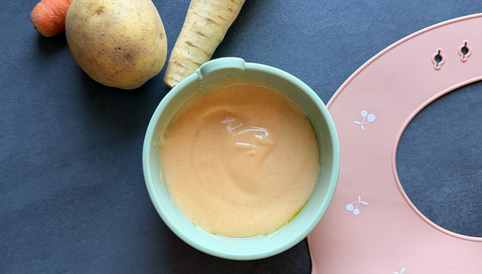 Gemüsepüree in einer grünen Schüssel, umgeben von einer Karotte, einem Stück Kartoffel, einer Pastinake und einem rosa Lätzchen.