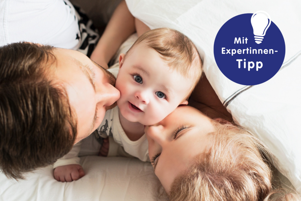 Eltern und Kleinkind liegen entspannt im Bett, umgeben von Decken. Das Kind lächelt glücklich zwischen den Eltern.