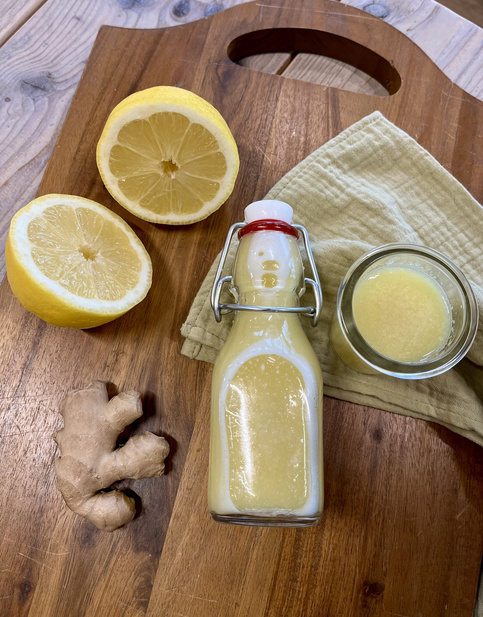 Hausgemachter Ingwer-Shot in einer Glasflasche, frische Zitrone und Ingwerknolle auf einem Holzbrett.