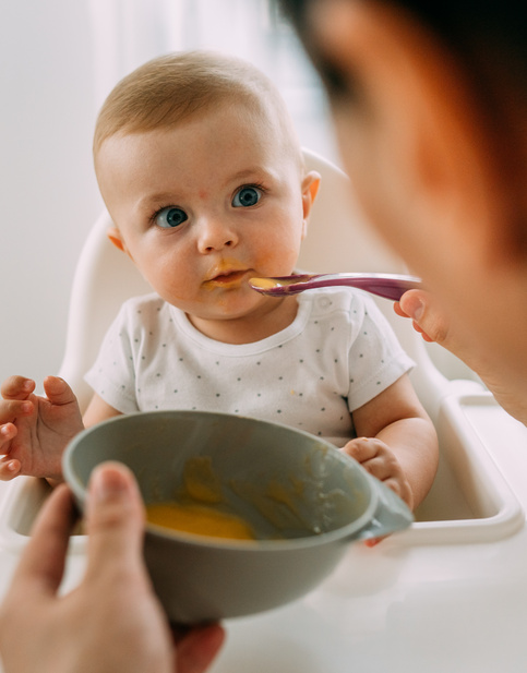 Baby in Hochstuhl wird mit Brei von Erwachsenen gefüttert.