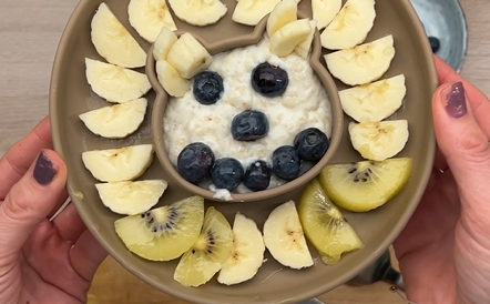Bowl mit Abendbrei, dekoriert mit Bananen, Heidelbeeren und Kiwi, gehalten von zwei Händen auf einem Holztisch.