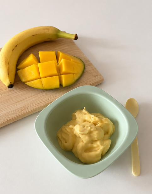 Fruchtiges Eis für babys und Kleinkinder aus Mango und Banane in einer grünen Schüssel, daneben frische, geschnittene Mango und eine Banane auf einem Holzbrett.