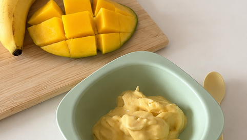 Fruchtiges Eis für babys und Kleinkinder aus Mango und Banane in einer grünen Schüssel, daneben frische, geschnittene Mango und eine Banane auf einem Holzbrett.