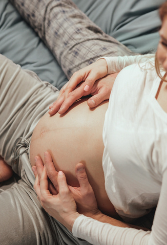 Paar lächelnd auf dem Bett, Hände auf Babybauch der schwangeren Frau ruhend.