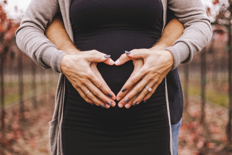 Hände formen ein Herz auf einem schwangeren Bauch im Freien.