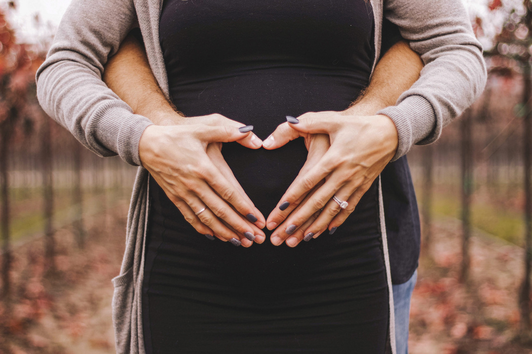 Hände formen ein Herz auf einem schwangeren Bauch im Freien.