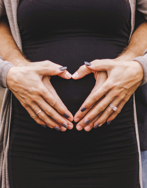 Mann umarmt Frau von hinten und legt seine Hände mit auf ihren Babybauch. Hände formen ein Herz auf einem Babybauch, symbolisieren Vorfreude und Zusammenhalt in der Schwangerschaft.