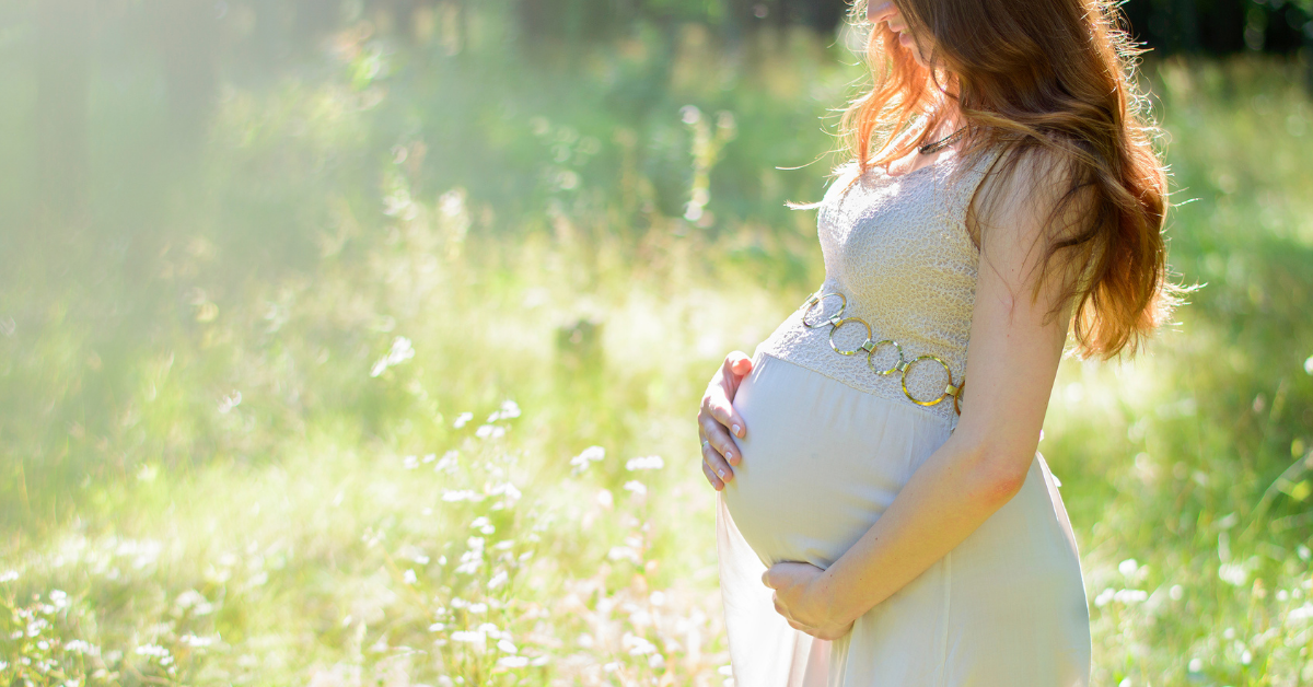 Schwangere Frau im Freien, die einen fließenden, pastellfarbenen Kleid trägt und sanft ihren Babybauch hält.